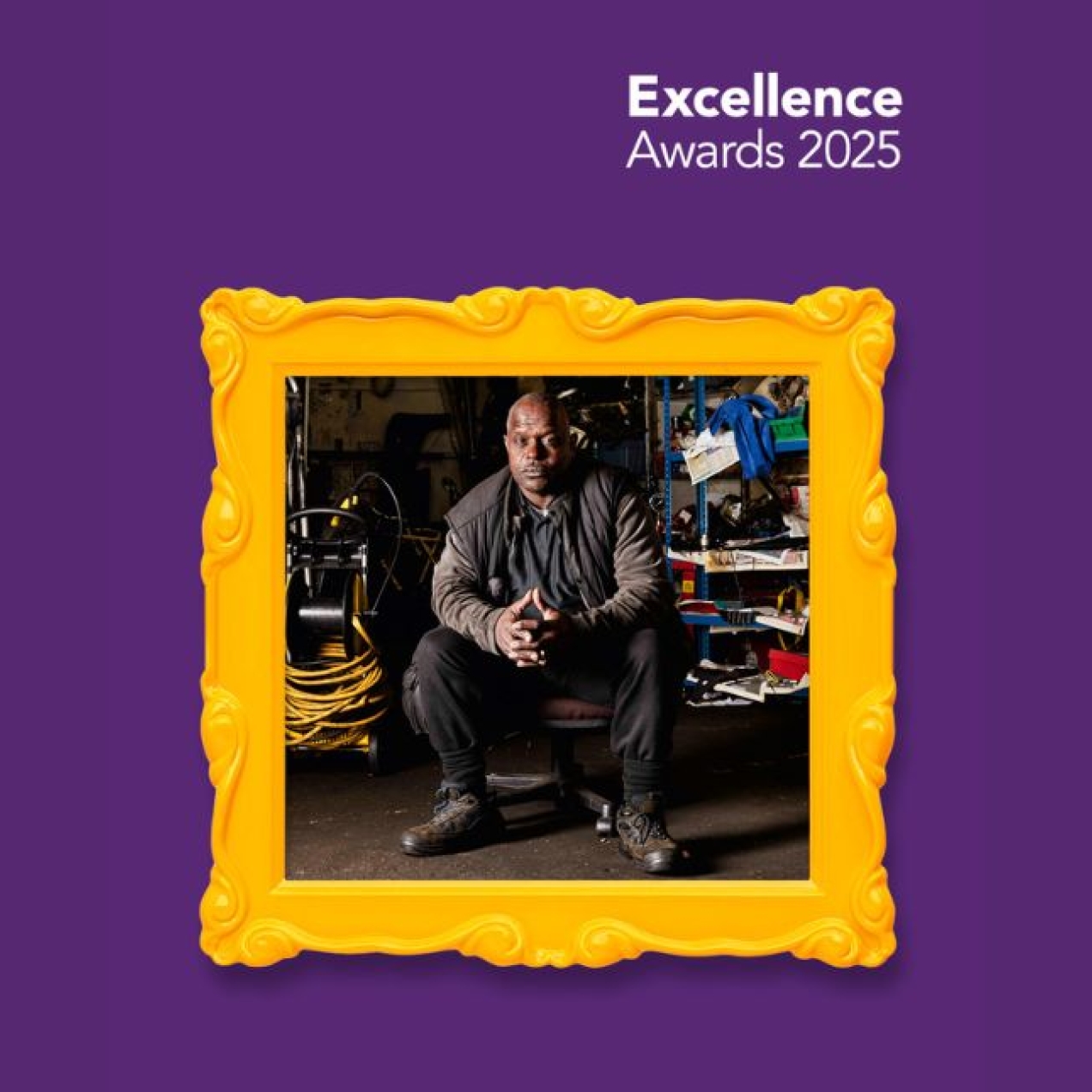 Superimposed on a purple background is a gold picture frame. Inside the frame is a photograph of a black man in black clothing sat in a workshop surrounded by tools and equipment.